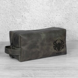 May include: A dark grey leather wash bag with a zip closure and a handle. The bag has a black spider emblem and the name "STEVE" printed beneath it. The rectangular bag is on a light-coloured surface.