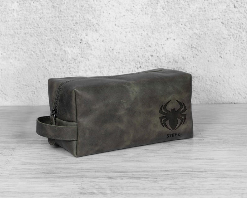 May include: A dark grey leather wash bag with a zip closure and a handle. The bag has a black spider emblem and the name "STEVE" printed beneath it. The rectangular bag is on a light-coloured surface.