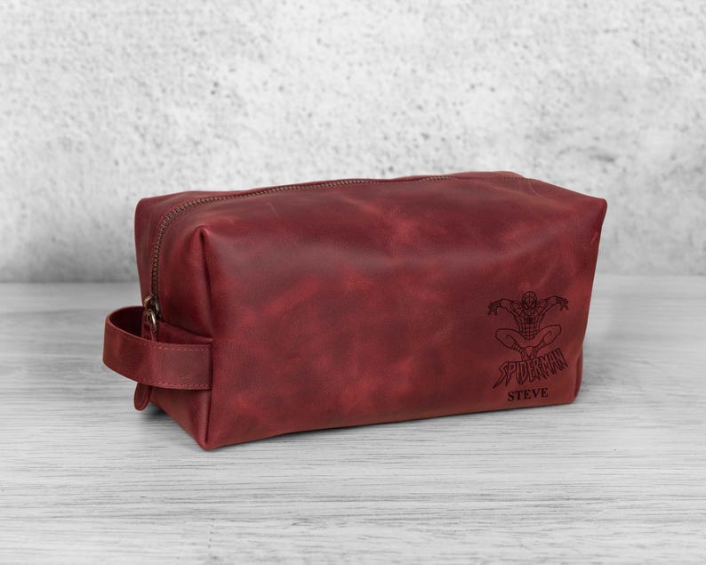 May include: A burgundy leather wash bag with a zip closure and a handle. The bag features a Spider-Man graphic and the name "STEVE" engraved on the front. The rectangular bag appears to be made of high-quality leather.