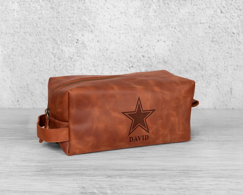 K&ouml;nnte beinhalten: Eine braune Leder-Kulturtasche mit Rei&szlig;verschluss und Griff. Die Tasche hat ein Stern-Design und den Namen "DAVID" auf der Vorderseite eingraviert. Die rechteckige Tasche steht auf einer hellen Oberfl&auml;che.