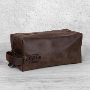 K&ouml;nnte beinhalten: Eine braune Leder-Kulturtasche mit Rei&szlig;verschluss und Griff. Auf der Tasche steht "San Antonio Spurs" und der Name "Thomas". Die Tasche ist rechteckig und hat einen Vintage-Look.