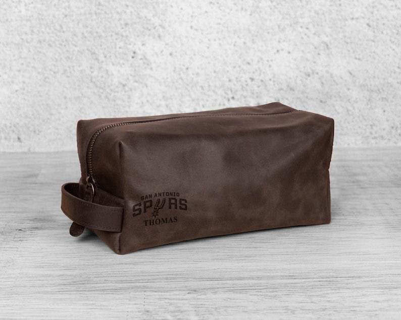 K&ouml;nnte beinhalten: Eine braune Leder-Kulturtasche mit Rei&szlig;verschluss und Griff. Auf der Tasche steht "San Antonio Spurs" und der Name "Thomas". Die Tasche ist rechteckig und hat einen Vintage-Look.