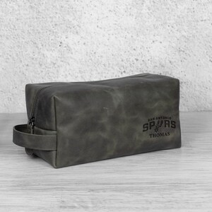 K&ouml;nnte beinhalten: Graue Leder-Kulturtasche mit Rei&szlig;verschluss und Griff. Die Tasche tr&auml;gt das Logo der San Antonio Spurs und den Namen "Thomas" auf der Seite.