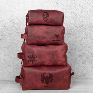 May include: Four stacked, burgundy leather wash bags. Each bag features a different Spider-Man emblem and a name. The bags vary in size and have a zip closure and a small handle on the side.