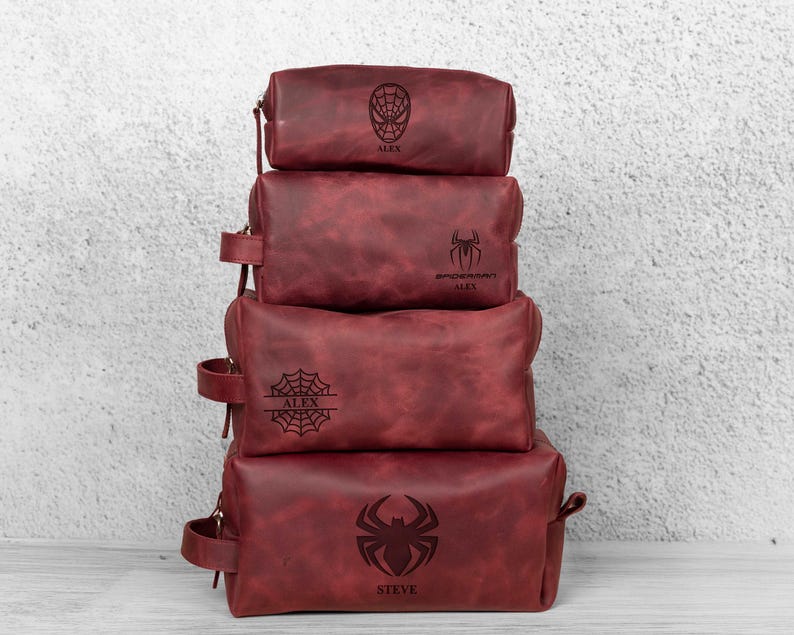 May include: Four stacked, burgundy leather wash bags. Each bag features a different Spider-Man emblem and a name. The bags vary in size and have a zip closure and a small handle on the side.
