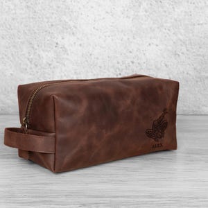 May include: A brown leather wash bag with a zip closure and a handle. The bag features a decorative design and the name "ALEX" engraved on the front. The rectangular bag is on a light-coloured surface.
