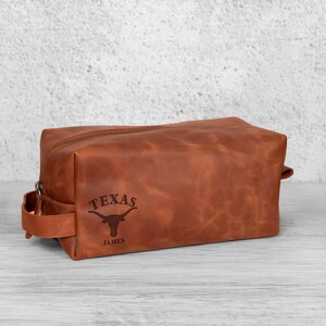 K&ouml;nnte beinhalten: Eine braune Leder-Kulturtasche mit Rei&szlig;verschluss und einem Griff an der Seite. Auf der Tasche sind das Wort "TEXAS" und ein Longhorn-Logo eingraviert, darunter der Name "JAMES".