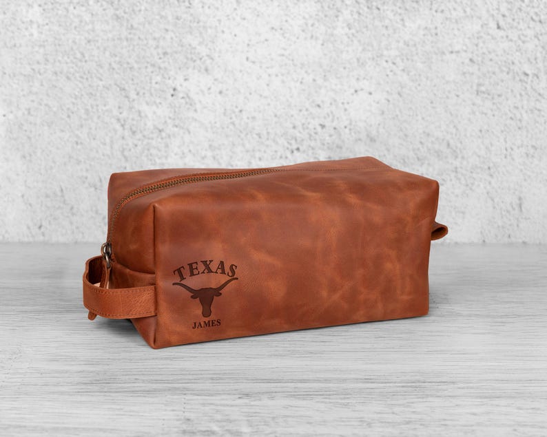 K&ouml;nnte beinhalten: Eine braune Leder-Kulturtasche mit Rei&szlig;verschluss und einem Griff an der Seite. Auf der Tasche sind das Wort "TEXAS" und ein Longhorn-Logo eingraviert, darunter der Name "JAMES".