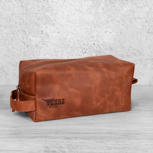 K&ouml;nnte beinhalten: Eine rechteckige braune Leder-Kulturtasche mit Rei&szlig;verschluss und seitlichem Griff. Auf der Tasche sind die Worte "TEXAS" und der Name "DAVID" aufgedruckt.
