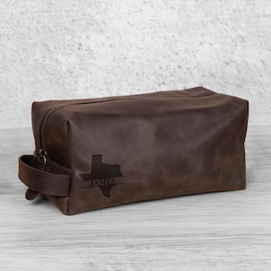 K&ouml;nnte beinhalten: Eine braune Leder-Kulturtasche mit Rei&szlig;verschluss und Griff. Die Tasche hat eine gepr&auml;gte Texas-Umrisslinie mit dem Namen "JAMES" darunter. Die rechteckige Tasche steht auf einer hellen Holzoberfl&auml;che.