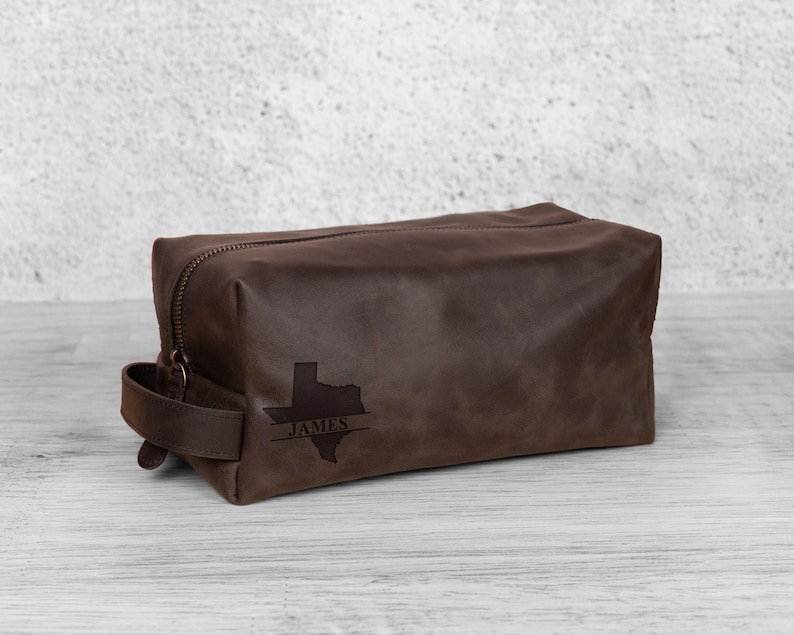 K&ouml;nnte beinhalten: Eine braune Leder-Kulturtasche mit Rei&szlig;verschluss und Griff. Die Tasche hat eine gepr&auml;gte Texas-Umrisslinie mit dem Namen "JAMES" darunter. Die rechteckige Tasche steht auf einer hellen Holzoberfl&auml;che.