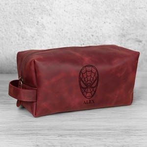 May include: A burgundy leather wash bag with a zip closure and a handle. The bag features a Spiderman mask design and the name "ALEX" below it. The rectangular bag is on a light-coloured wooden surface.
