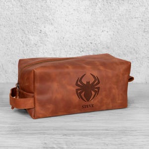 May include: A brown leather wash bag with a zip closure and a handle. It has a spider emblem and the name "STEVE" engraved on the front. The rectangular bag is on a light-coloured wooden surface. The bag is designed for toiletries.