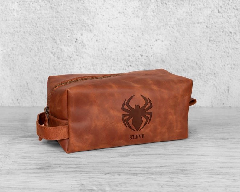 May include: A brown leather wash bag with a zip closure and a handle. It has a spider emblem and the name "STEVE" engraved on the front. The rectangular bag is on a light-coloured wooden surface. The bag is designed for toiletries.