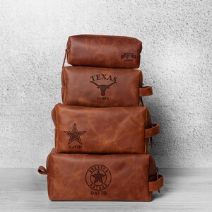 K&ouml;nnte beinhalten: Vier gestapelte braune Leder-Kulturtaschen. Jede Tasche hat ein anderes Design: Spurs, Texas Longhorn, ein Stern und das Houston Astros-Logo. Jede Tasche hat einen Rei&szlig;verschluss und einen kleinen Griff an der Seite. Die Taschen sind mit Namen personalisiert.