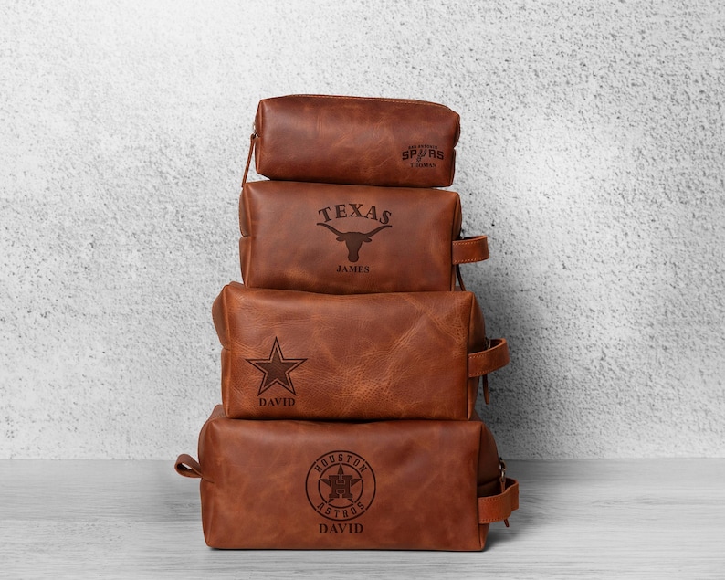 K&ouml;nnte beinhalten: Vier gestapelte braune Leder-Kulturtaschen. Jede Tasche hat ein anderes Design: Spurs, Texas Longhorn, ein Stern und das Houston Astros-Logo. Jede Tasche hat einen Rei&szlig;verschluss und einen kleinen Griff an der Seite. Die Taschen sind mit Namen personalisiert.