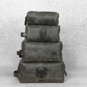 May include: Four grey leather wash bags stacked on top of each other. Each bag has a different Spider-Man design, including a spider web, a mask, and a spider symbol. The names "ALEX" and "STEVE" are visible.
