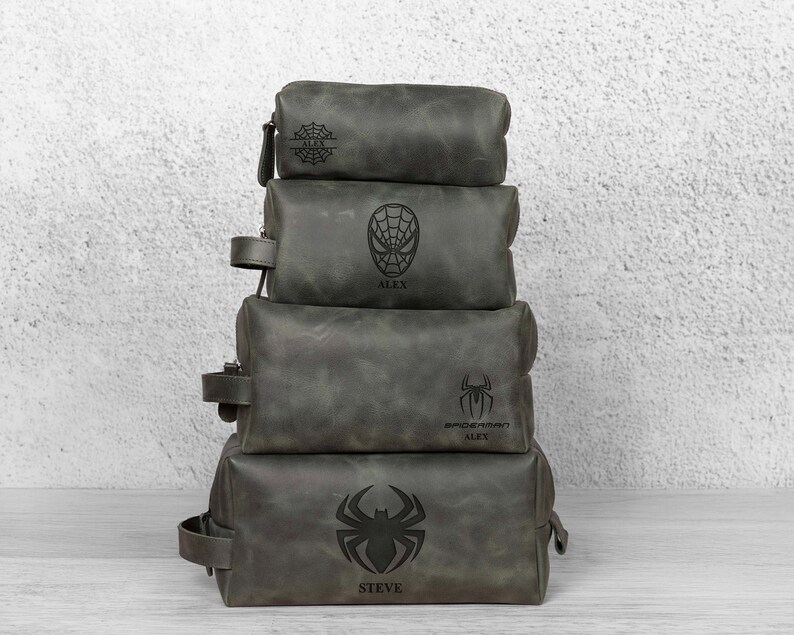 May include: Four grey leather wash bags stacked on top of each other. Each bag has a different Spider-Man design, including a spider web, a mask, and a spider symbol. The names "ALEX" and "STEVE" are visible.