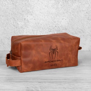 May include: A brown leather wash bag with a zip closure and a handle on the side. The bag features a Spiderman logo and the name "ALEX" engraved on the front. The rectangular bag sits on a light-coloured surface.