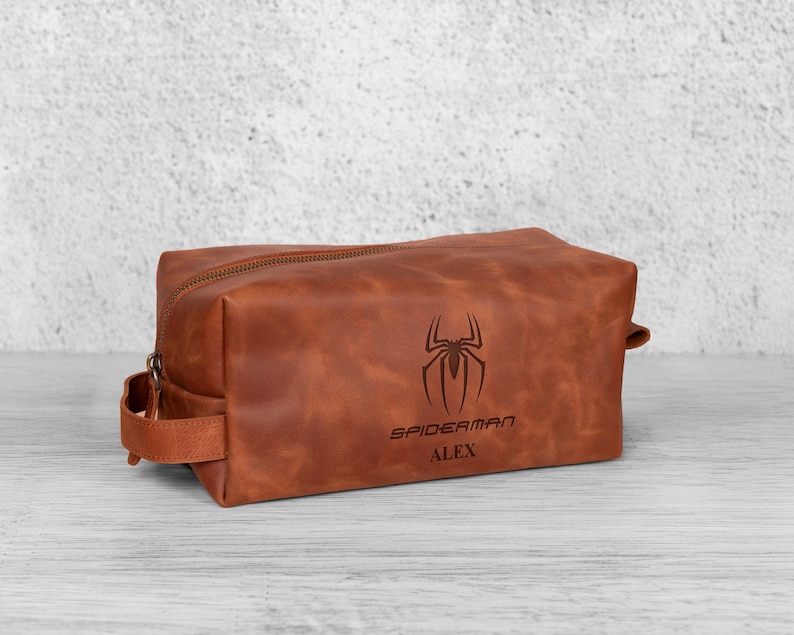 May include: A brown leather wash bag with a zip closure and a handle on the side. The bag features a Spiderman logo and the name "ALEX" engraved on the front. The rectangular bag sits on a light-coloured surface.