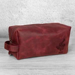 May include: A burgundy leather wash bag with a zip closure and a handle. The bag features a Spiderman graphic and the name "ALEX" engraved on the front. The bag is rectangular and sits on a light wood surface.