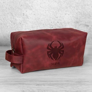 May include: A burgundy leather wash bag with a zip closure and a handle. The bag features a debossed spider emblem and the name "STEVE" below it. The rectangular bag sits on a light-coloured wooden surface.