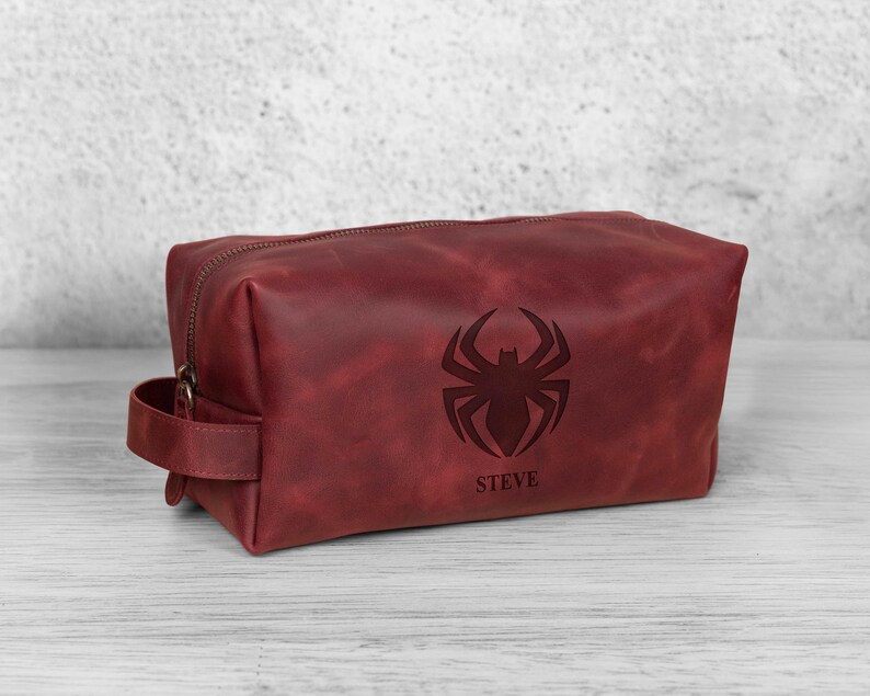 May include: A burgundy leather wash bag with a zip closure and a handle. The bag features a debossed spider emblem and the name "STEVE" below it. The rectangular bag sits on a light-coloured wooden surface.