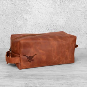K&ouml;nnte beinhalten: Eine braune Leder-Kulturtasche mit Rei&szlig;verschluss und Griff. Auf der Tasche ist ein Longhorn-Logo und der Name "THOMAS" eingraviert. Die rechteckige Tasche ist aus hochwertigem Leder gefertigt und f&uuml;r Toilettenartikel gedacht.