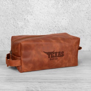 K&ouml;nnte beinhalten: Eine braune Leder-Kulturtasche mit Rei&szlig;verschluss und Griff. Auf der Tasche ist das Wort "TEXAS" und ein Longhorn-Logo auf der Vorderseite eingepr&auml;gt, darunter der Name "DAVID".
