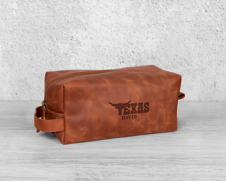 K&ouml;nnte beinhalten: Eine braune Leder-Kulturtasche mit Rei&szlig;verschluss und Griff. Auf der Tasche ist das Wort "TEXAS" und ein Longhorn-Logo auf der Vorderseite eingepr&auml;gt, darunter der Name "DAVID".