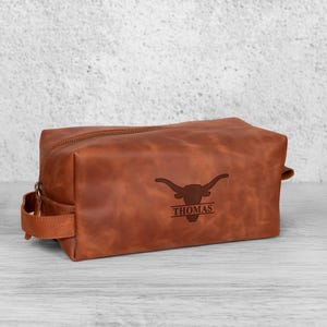K&ouml;nnte beinhalten: Eine braune Leder-Kulturtasche mit Rei&szlig;verschluss und Griff an der Seite. Die Tasche hat ein Longhorn-Logo und den Namen "THOMAS" auf der Vorderseite eingraviert.