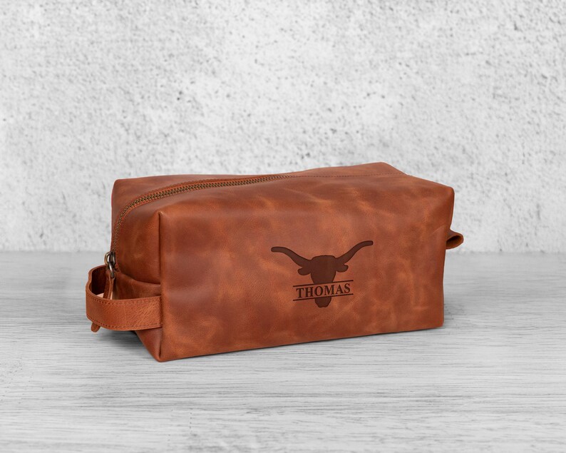 K&ouml;nnte beinhalten: Eine braune Leder-Kulturtasche mit Rei&szlig;verschluss und Griff an der Seite. Die Tasche hat ein Longhorn-Logo und den Namen "THOMAS" auf der Vorderseite eingraviert.