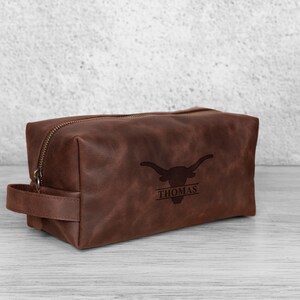 K&ouml;nnte beinhalten: Eine braune Leder-Kulturtasche mit Rei&szlig;verschluss und Griff. Die Tasche zeigt ein Longhorn-Logo und den Namen "THOMAS" darunter. Die rechteckige Tasche steht auf einer hellen Oberfl&auml;che.