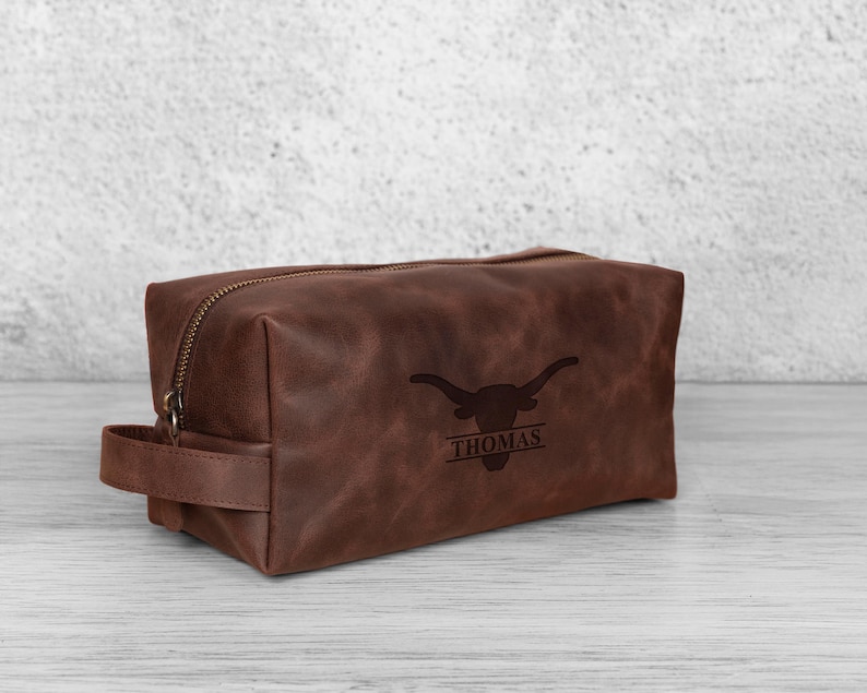 K&ouml;nnte beinhalten: Eine braune Leder-Kulturtasche mit Rei&szlig;verschluss und Griff. Die Tasche zeigt ein Longhorn-Logo und den Namen "THOMAS" darunter. Die rechteckige Tasche steht auf einer hellen Oberfl&auml;che.