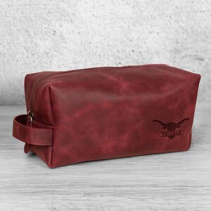 K&ouml;nnte beinhalten: Eine bordeauxfarbene Leder-Kulturtasche mit Rei&szlig;verschluss und Griff. Die Tasche hat ein Longhorn-Logo und den Namen "Thomas" auf der Vorderseite eingraviert. Die Tasche ist rechteckig und steht auf einer hellen Holzoberfl&auml;che.