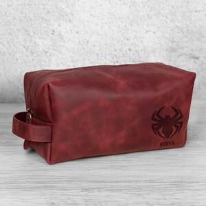 May include: A burgundy leather wash bag with a zip closure and a handle. The bag features a spider emblem and the name "STEVE" engraved on the front. The bag is rectangular and sits on a light-coloured wooden surface.