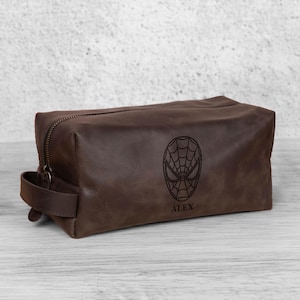 May include: A brown leather wash bag with a zip closure and a handle. The bag features a Spiderman mask design and the name "ALEX" engraved on the front. The bag is rectangular and appears to be made of high-quality leather.