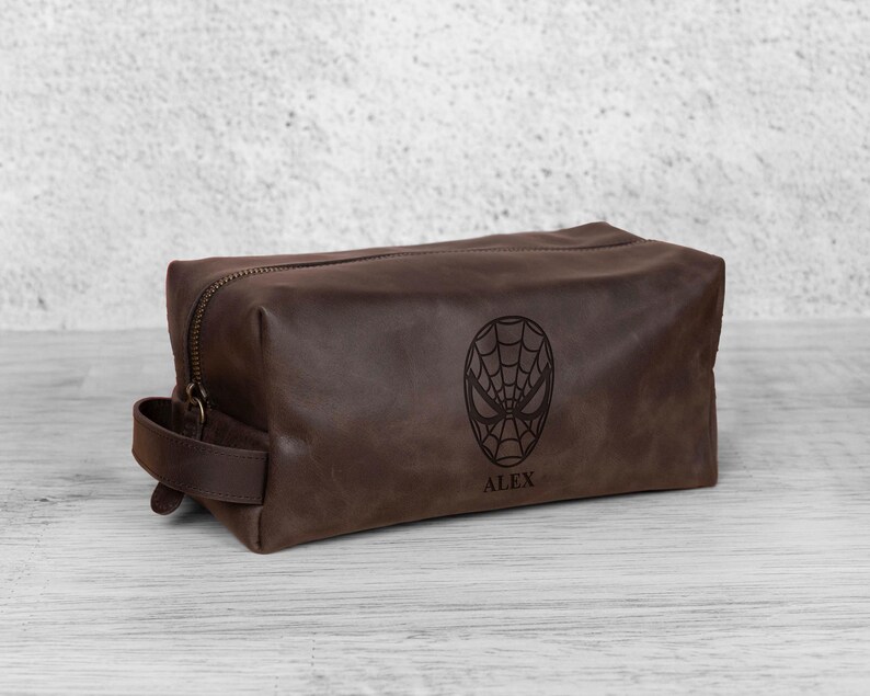 May include: A brown leather wash bag with a zip closure and a handle. The bag features a Spiderman mask design and the name "ALEX" engraved on the front. The bag is rectangular and appears to be made of high-quality leather.