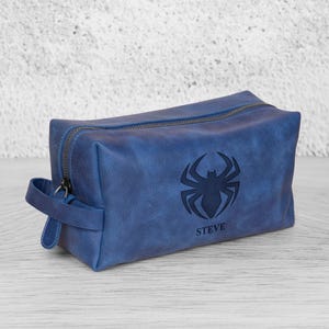 May include: A blue leather wash bag with a zip closure and a handle. The bag has a black spider emblem and the name "STEVE" beneath it. The rectangular bag appears to be made of a durable material, suitable for travel.