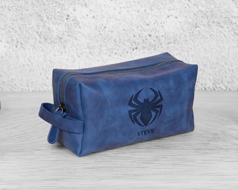 May include: A blue leather wash bag with a zip closure and a handle. The bag has a black spider emblem and the name "STEVE" beneath it. The rectangular bag appears to be made of a durable material, suitable for travel.