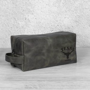 K&ouml;nnte beinhalten: Eine graue Leder-Kulturtasche mit Rei&szlig;verschluss und Griff. Auf der Tasche steht das Wort "TEXAS" und ein Longhorn-Logo, darunter der Name "JAMES". Die rechteckige Tasche steht auf einer hellen Oberfl&auml;che.