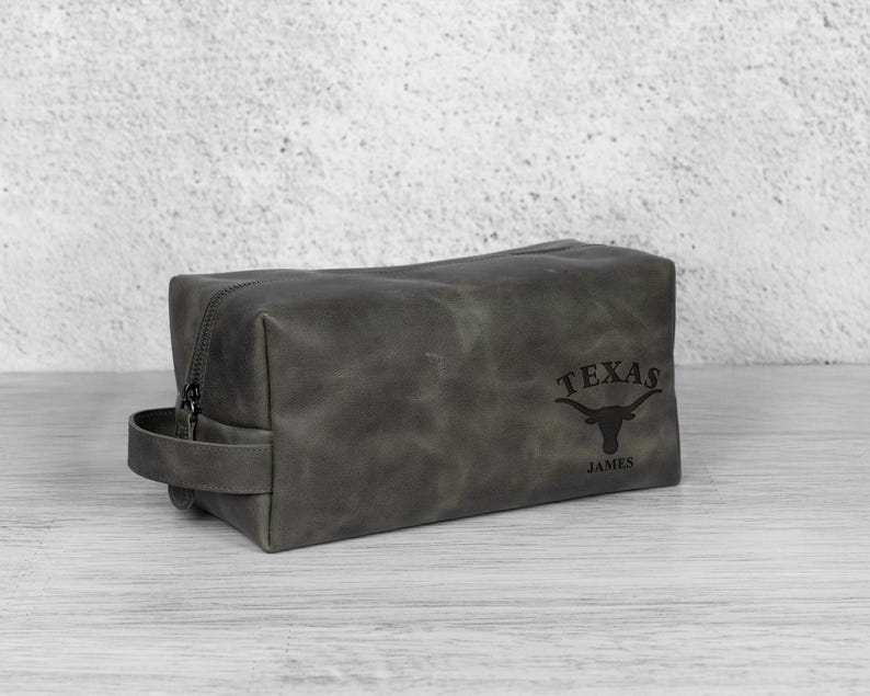 K&ouml;nnte beinhalten: Eine graue Leder-Kulturtasche mit Rei&szlig;verschluss und Griff. Auf der Tasche steht das Wort "TEXAS" und ein Longhorn-Logo, darunter der Name "JAMES". Die rechteckige Tasche steht auf einer hellen Oberfl&auml;che.