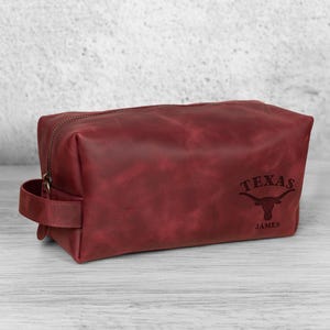 K&ouml;nnte beinhalten: Eine bordeauxfarbene Kulturtasche aus Leder mit Rei&szlig;verschluss und Griff. Auf der Tasche steht das Wort "TEXAS" &uuml;ber einem Longhorn-Logo und der Name "JAMES" darunter.
