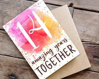 May include: A greeting card with the number 14 and the words "amazing years TOGETHER" on a watercolor background of pink, red, and yellow. A brown envelope, a cup of coffee with a heart design, and a pen are also on the wooden surface.