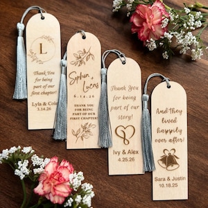 May include: Wooden bookmarks with gray tassels. Each bookmark has a unique design and text, including names and dates. Floral accents and a heart symbol are also present. The bookmarks are arranged on a wooden surface.