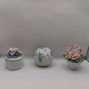Könnte beinhalten: Drei dekorative Keramikartikel: eine sechseckige Schatulle mit Blumen, ein runder Ornament mit blauem Blumendesign und ein Korb mit einer Rose. Die Artikel sind weiß mit pastellfarbenen floralen Akzenten.