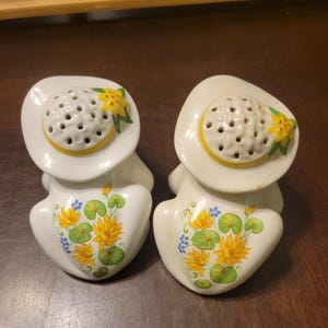 May include: A pair of vintage ceramic salt and pepper shakers. Each shaker is white with a hat-shaped top featuring multiple holes. The hats have a yellow band and a yellow flower with green leaves. The bodies are decorated with yellow flowers and green leaves.