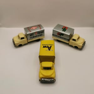 May include: Three vintage toy trucks in yellow and silver. One truck has a yellow cab and a yellow container with the text "AW LIVESTOCK". The other two trucks have yellow cabs and silver containers with the text "ABC Freight Fowarding" and "Consolidated Freight Ways".
