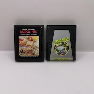 Peut inclure: Deux cartouches de jeux Atari vintage : "Dodge 'Em" et "Frogger". La cartouche "Dodge 'Em" a une étiquette colorée avec des graphiques et du texte. La cartouche "Frogger" présente un graphisme de grenouille. Les deux sont en plastique noir.