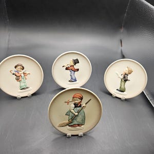 Könnte beinhalten: Ein Set aus vier runden Wandtellern aus Keramik mit erhabenen, handbemalten Figuren. Ein Teller zeigt ein Kind mit einem Besen, während die anderen Kinder beim Musizieren oder mit verschiedenen Gegenständen in gedeckten Erdtönen darstellen.